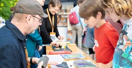 Zwei Kinder stehen vor einem Tisch und schauen sich Ausstellungsstücke an, während ein Mann ihnen etwas erklärt