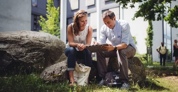 Zwei Studierende sitzen auf dem Campus und schauen auf ein Tablet.