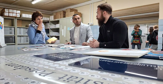 Drei Personen sind um einen Tisch versammelt auf dem Druckerzeugnisse ausgelegt sind.