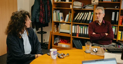Prof. Dr. Christoph Schroeder und Dorotheé Steinbock im Gespräch an einem Tisch in einem Büro sitzend. Im Hintergrund in einem Regal Bücher, Aktenordner und Tassen.