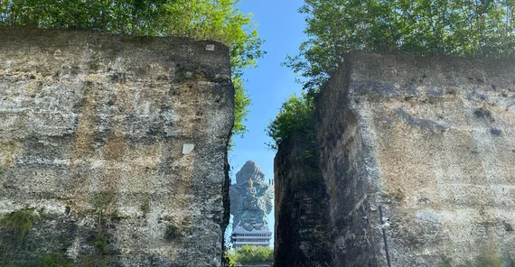 Die Garuda-Wisnu-Kencana-Statue ist in der Ferne im Hintergrund zu erkennen. Sie ist sehr groß und thront auf der gesamten Dachfläche eines großen Gebäudes. Die Statue ist durch den Spalt zwischen einer Felsformation mit sekrechten Wänden zu erkennen.