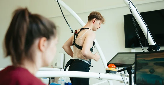 Eine junge Frau schaut auf einen Bildschirm. Hinter ihr läuft ein junger Mann auf einem Laufband. Er ist verkabelt und mit einem Seil gesichert.