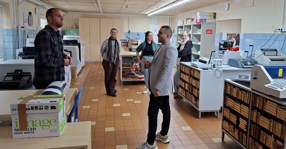 Zwei Personen im Vordergrund unterhalten sich in einem großen Raum. Im Hintergrund stehen weitere Personen.