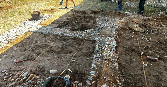 Bild einer Ausgrabungsstelle mit Steinfragmenten, die den Grundriss eines Hauses bilden.