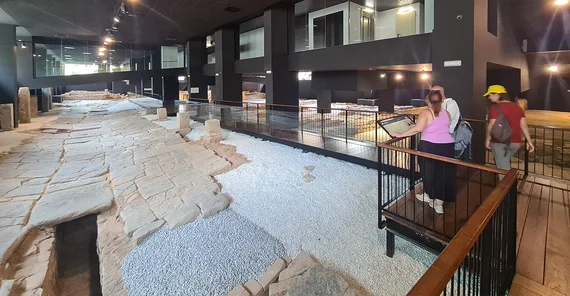 Ein freigelegter römischer Straßenzug unter dem Bodenniveau der heutigen Stadt. Der Straßenzug ist überdacht und von künstlichem Licht beschienen. Menschen können ihn nicht betreten, für sie sind Plattformen mit Absperrungen installiert.