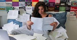 Junge Frau liest in einem Heft auf einem Bett voller Papiere. Foto: Sophia Pakzad