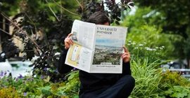 Auf dem Foto sieht man einen schwarz gekleideten Mann, der die neueste Ausgabe der Beilage im Park liest.