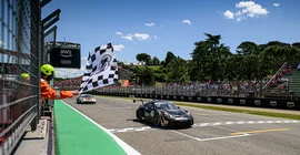 Ein schwarzer Porsche 911 mit goldenen Details fährt über die Ziellinie. Links schwenkt jemand mit Helm und Kopfhörern eine schwarz-weiß-karierte Zielflagge. Im Hintergrund sieht man volle Zuschauerränge und Bäume. Der Himmel ist blau mit einzelnen, weißen Wolken.