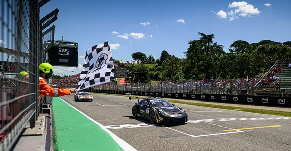 Ein schwarzer Porsche 911 mit goldenen Details fährt über die Ziellinie. Links schwenkt jemand mit Helm und Kopfhörern eine schwarz-weiß-karierte Zielflagge. Im Hintergrund sieht man volle Zuschauerränge und Bäume. Der Himmel ist blau mit einzelnen, weißen Wolken.