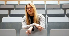 Frau sitzt im Hörsaal in Sizreihen und blickt in die Kamera.