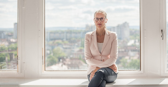 Frau sitzt vor einem großen Fenster und schaut in die Kamera