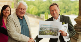 Zwei Männer schütteln einander die Hand, einer hält einen Ausdruck eines Bauentwurfs in die Kamera