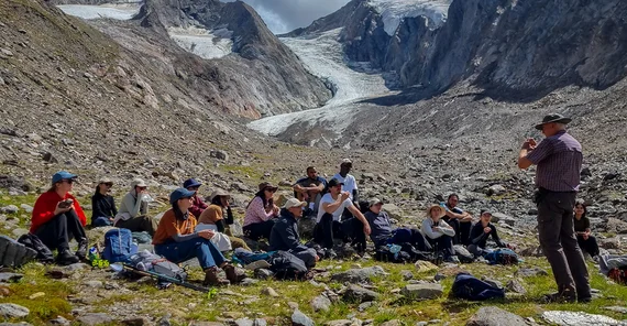 People sitting in front of an impressive mountain scape while one of them is standing in front of them explaining.