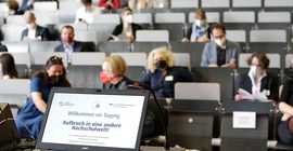Das Bild zeigt einen Laptop an einem Rednerpult mit dem Tagungstitel "Willkommen zur Tagung. Aufbruch in eine andere Hochschulwelt!" und im Hintergrund das Publikum. Foto: Sandra Scholz
