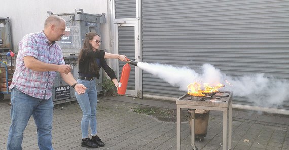 Brandschutzübung im Rahmen der Erstunterweisung in Arbeits- und Umweltschutz. Eine Person löscht unter Anleitung des Leiters des Sicherheitswesens der UP einen kontrollierten Brand in einem Topf mit einem Feuerlöscher.