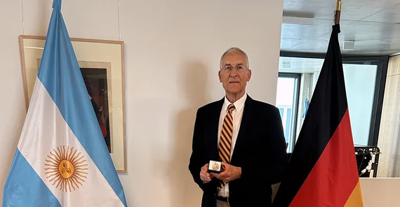 Ein Mann steht zwischen einer argentinischen und einer deutschen Flagge und präsentiert eine Auszeichnung
