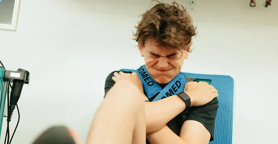 Ein junger Mann mit dunklen Haaren sitzt mit vor der Brust gekreuzten Armen auf einem Sitz. Er drückt mit einem Bein ein Gewicht weg, wobei er die Backen aufbläst und die Augen zukneift.