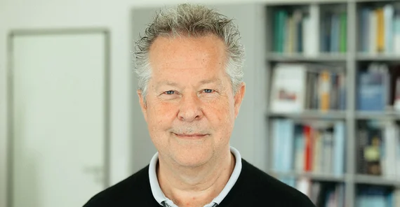 Ein Mann mit grauen Haaren in einem schwarzen Pullover. Im Hintergrund sieht man unscharf ein Bücherregal.