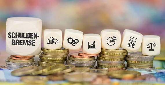 Symbolbild: Auf mehrern Stapel Hartgeld liegen diverse Spielwürfel. Auf ihnen sind u.a. Symbole für Berechnungen und Statistiken zu sehen. Auf dem größten Würfel ganz links steht "Schuldenbremse".