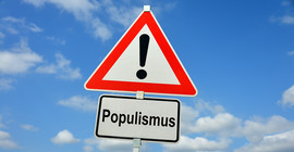 Dreieckiges Warnschild mit Ausrufezeichen, darunter Hinweisschild mit dem Begriff "Populismus". Beide vor blauem Himmel mit Wolken.