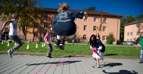 Kinder springen über ein langes Seil, gedreht von einem Mädchen in der Mitte. Im Hintegrund Uni-Gebäude.