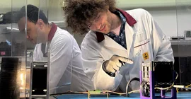 Szene im Labor: Eine Personen mit weißem Kittel deutet mit einer gummibehandschuhten Hand auf ein Gerät.