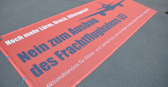 Rotes Protestbanner liegt auf dem Boden mit den Worten "Noch mehr Lärm, Dreck und Klimagase? Nein zum Ausbau des Frachtflughafens (E) Aktionsbündnis für Klima- und Lärmschutz und sofortigen Ausbaustopp am Flughafen Leizig/Halle"