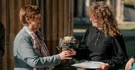 Eine Frau überreicht einer anderen eine Urkunde und einen Blumenstrauß