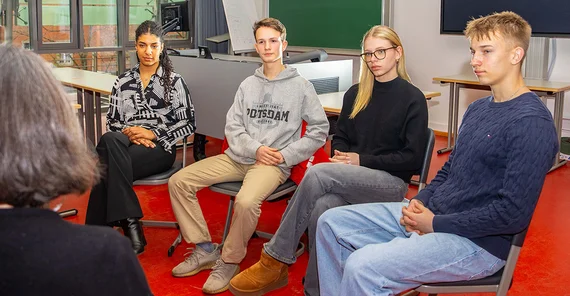 Vier Schüler*innen sitzen auf Stühlen und hören einen Vortrag.
