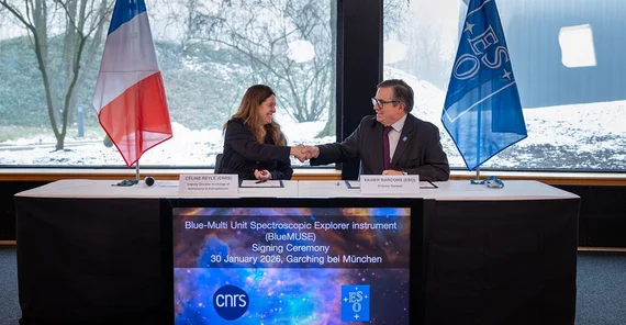 Zwei Personen schütteln sich im offiziellen Rahmen die Hände. Hinter ihnen einmal die französische Flagge und die ESO-Flagge.
