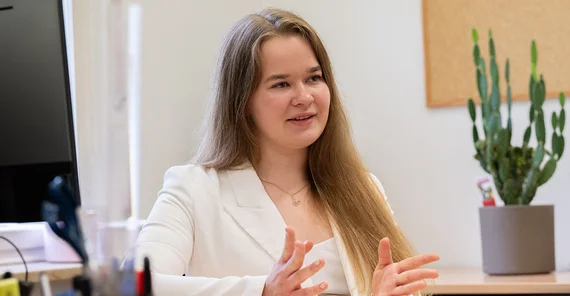 Eine junge Frau ein weiß erklärt etwas, im Hintergrund stehen ein Kaktus und Büromaterialien.